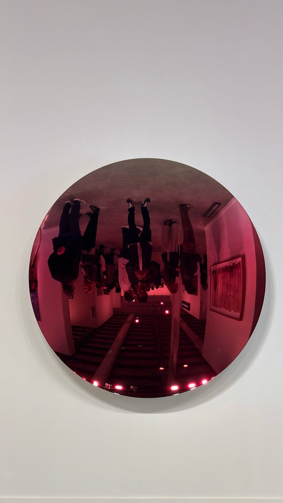 Espelho côncavo vermelho de Anish Kapoor, numa parede branca, refletindo de cabeça para baixo os visitantes e o interior da galeria