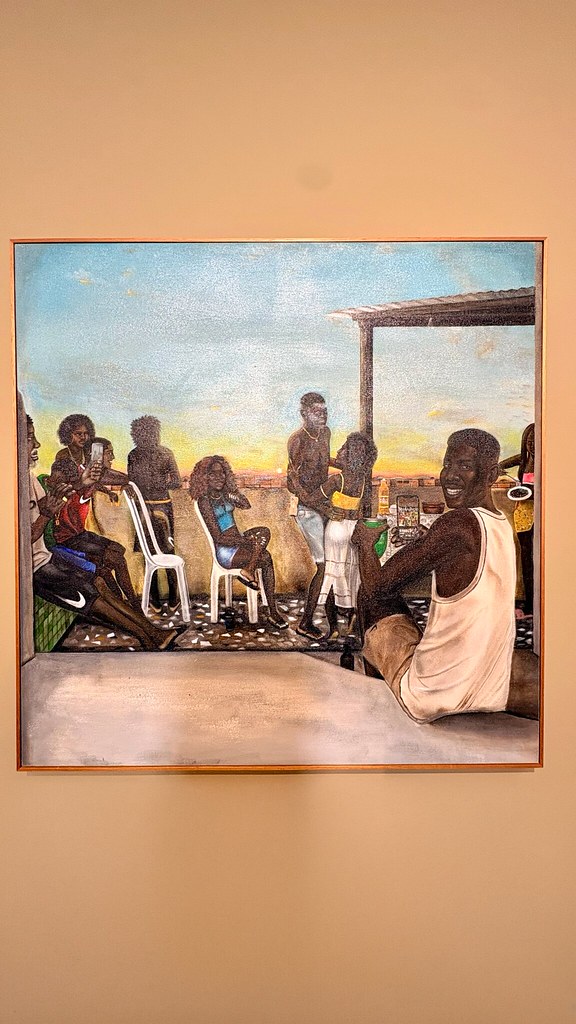 Pintura Feijoada de domingo, de Jota: pessoas reunidas num terraço ao fim da tarde, com cadeiras brancas de plástico e, à direita, um homem de regata branca sorrindo